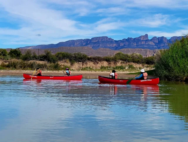 Three red canoes with people paddling on a calm river, under a clear blue sky and mountains in the background.