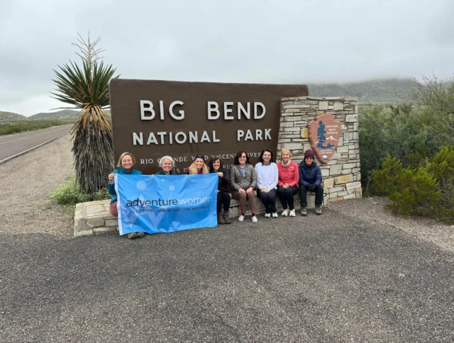 A group of seven women sitting beside a sign for Big Bend National Park, holding a blue "Adventure Women" banner. The setting is outdoors with cloudy skies.