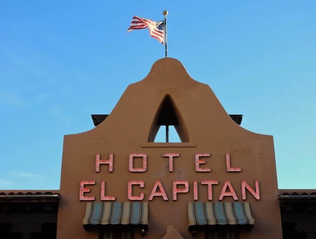 Hotel El Capitan's building facade with a neon sign and an American flag on top against a clear blue sky.