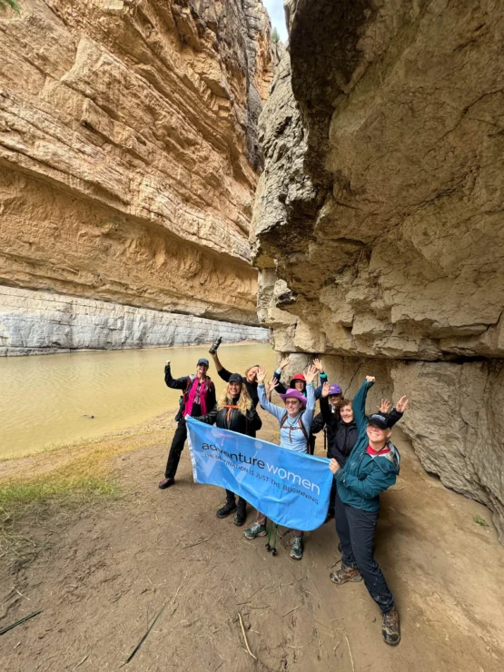 Group of people holding an "Adventure Women" banner, posing joyfully in a canyon near a river.