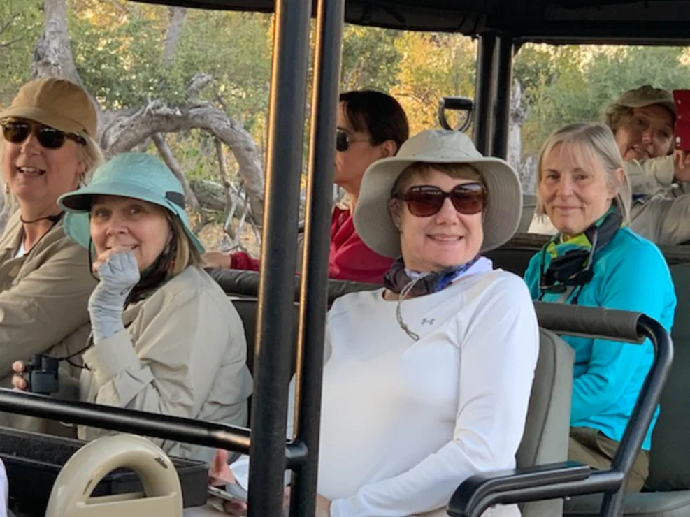 A group of people wearing hats and sunglasses sit in an open safari vehicle, smiling at the camera, surrounded by trees.