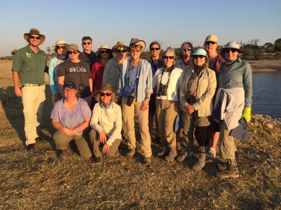 A group of people, wearing hats and outdoor clothing, stand together on a grassy area near a body of water under a clear sky. Some hold cameras.