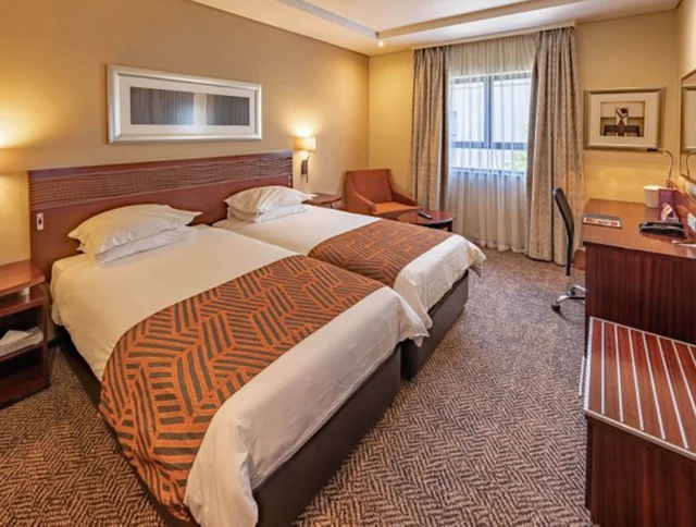 A hotel room with two single beds, decorative bed runners, a chair, desk, and window with curtains. Walls are light-colored with framed art above the beds.