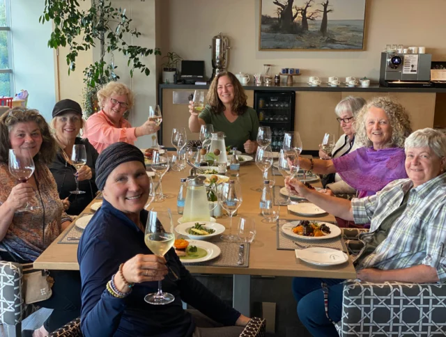 A group of smiling people sitting around a dining table, holding wine glasses for a toast, with plates of food in front of them.