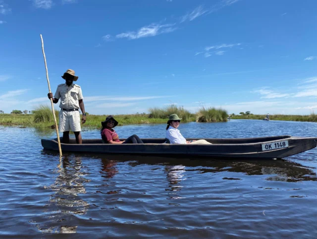 Three people in a small boat on a calm body of water, with one person standing and using a wooden pole. The sky is clear and blue.