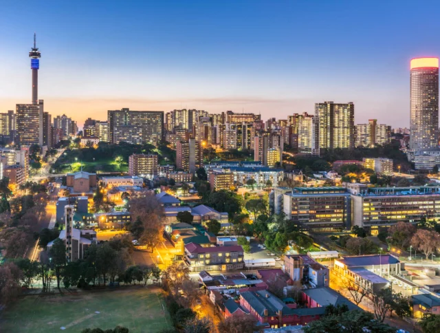 Aerial view of a cityscape at dusk with illuminated buildings, a prominent tower, and a glowing cylindrical skyscraper under a clear sky.