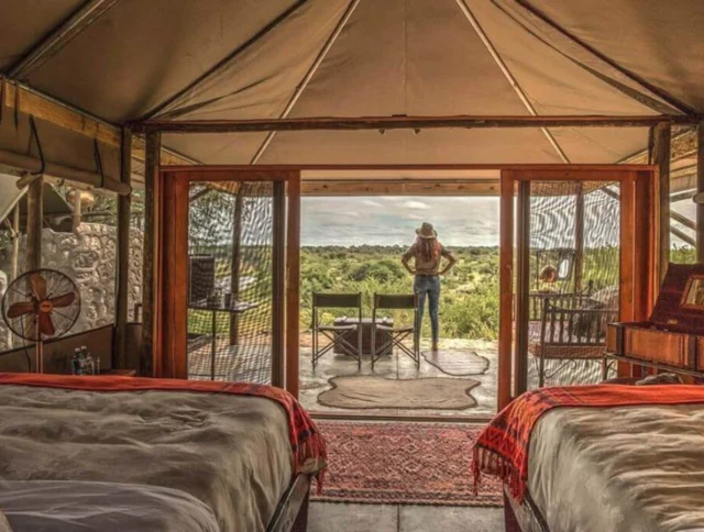 Interior of a luxury tent with two beds and an open entrance. A person stands outside, overlooking a scenic landscape.