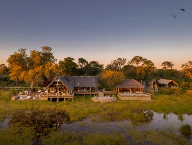 Aerial view of a lodge with thatched roofs surrounded by trees and a wetland area at sunset. Birds are flying in the sky.