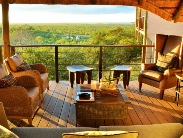 A cozy balcony with wicker and leather chairs overlooks a lush green landscape and distant water under a thatched roof, bathed in warm sunlight.