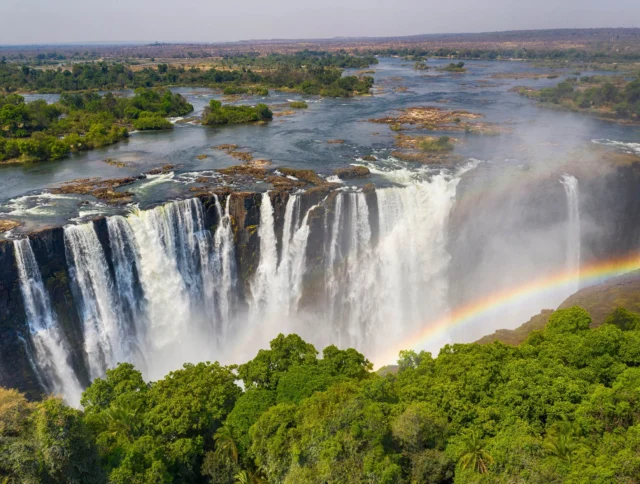 Aerial view of a large waterfall with a rainbow spanning across the mist, surrounded by lush green forest and flowing river.