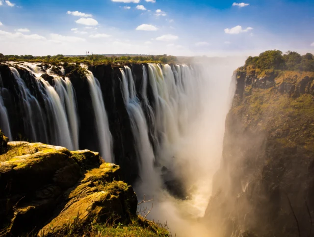 A wide waterfall flows over a cliff into a deep gorge surrounded by lush greenery under a partly cloudy sky.