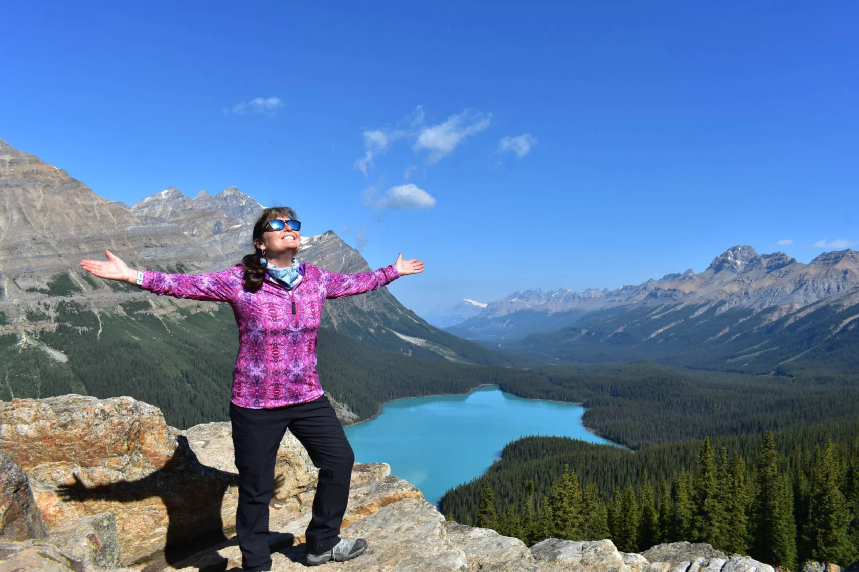 A person in a pink jacket stands on a rocky ledge with arms outstretched, overlooking a turquoise lake surrounded by mountains and forest under a clear blue sky.