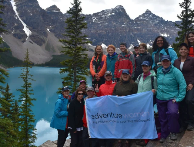 A group of women poses with an "Adventure Women" banner in front of a scenic mountain lake.