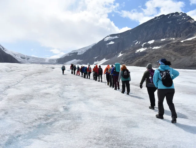 A group of people hikes across a glacier under a partly cloudy sky, surrounded by mountains.