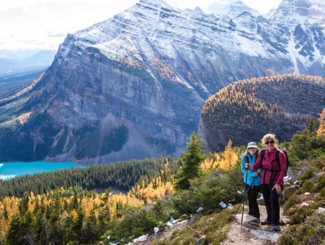 Two hikers with trekking poles stand on a mountain trail overlooking a turquoise lake and snow-capped peaks under a clear sky.