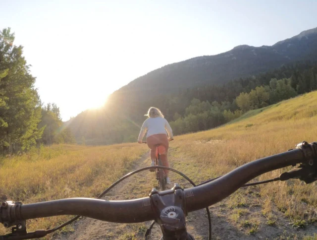 View from a bicycle following another cyclist on a grassy trail at sunrise, surrounded by trees and hills.