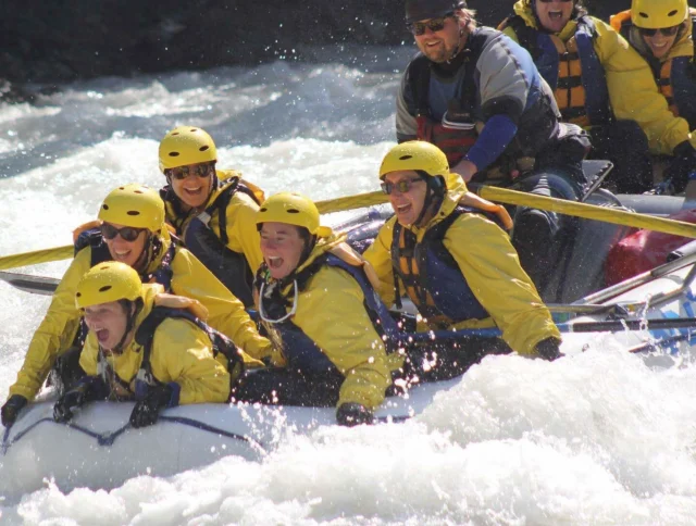 A group of people wearing helmets and life jackets navigate a whitewater rafting trip, surrounded by rushing water.