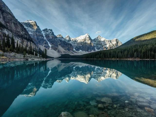 Snow-capped mountains reflected in a clear blue lake, surrounded by dense green pine forests under a sky streaked with clouds.