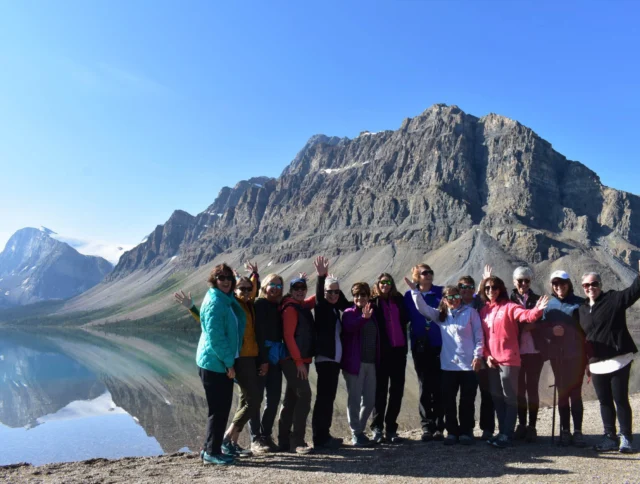 A group of people stand on a lakeshore, waving with a large mountain in the background under a clear blue sky.