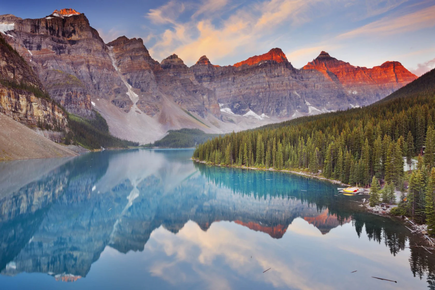 A serene mountain lake scene with clear blue water reflecting surrounding pine trees and peaks under a colorful sunset sky. Canoes are visible near a forested shore.