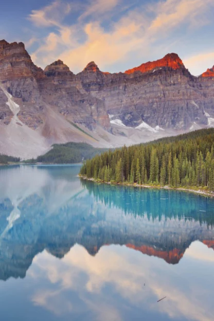 A serene mountain lake scene with clear blue water reflecting surrounding pine trees and peaks under a colorful sunset sky. Canoes are visible near a forested shore.