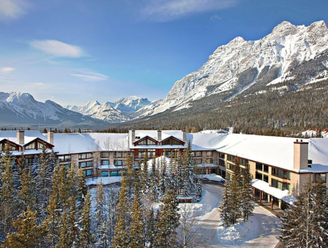 Snow-covered mountain resort with chalet-style buildings surrounded by evergreen trees. Majestic mountains and clear blue sky in the background.