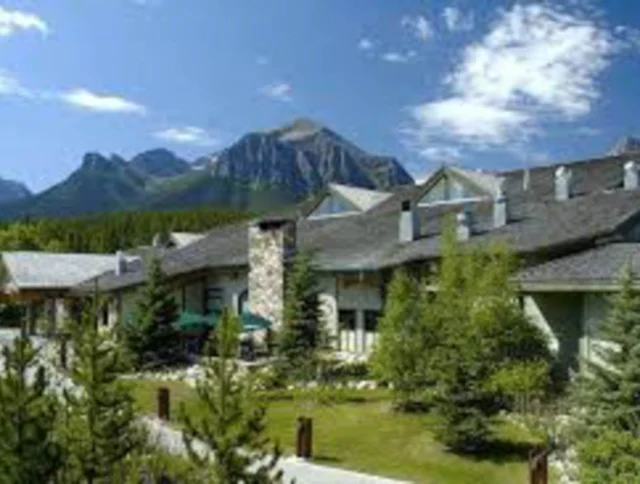 A building with a sloped roof surrounded by trees, set against a mountain backdrop under a partly cloudy sky.