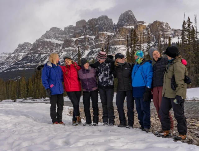 A group of people dressed in winter clothing stand together on snow-covered ground with a mountain range in the background.