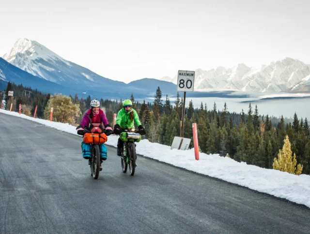 Two cyclists, wearing bright outdoor gear, ride bikes on a snow-lined road with a scenic mountain view and an 80 km/h speed limit sign.