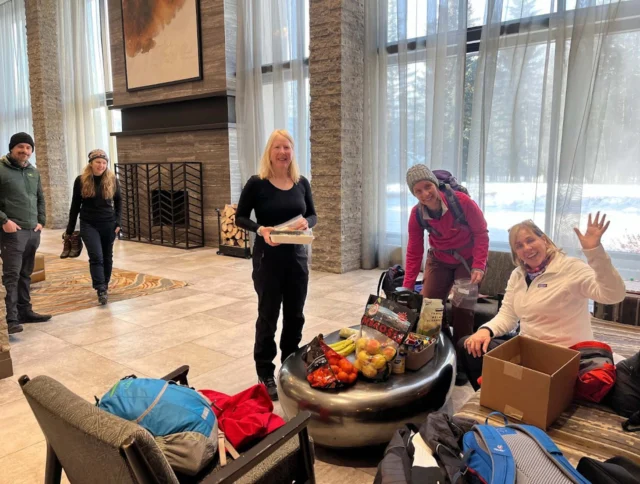 A group of people in a hotel lobby sorts through bags and snacks on a table. One person is waving, and there are backpacks and winter clothing.