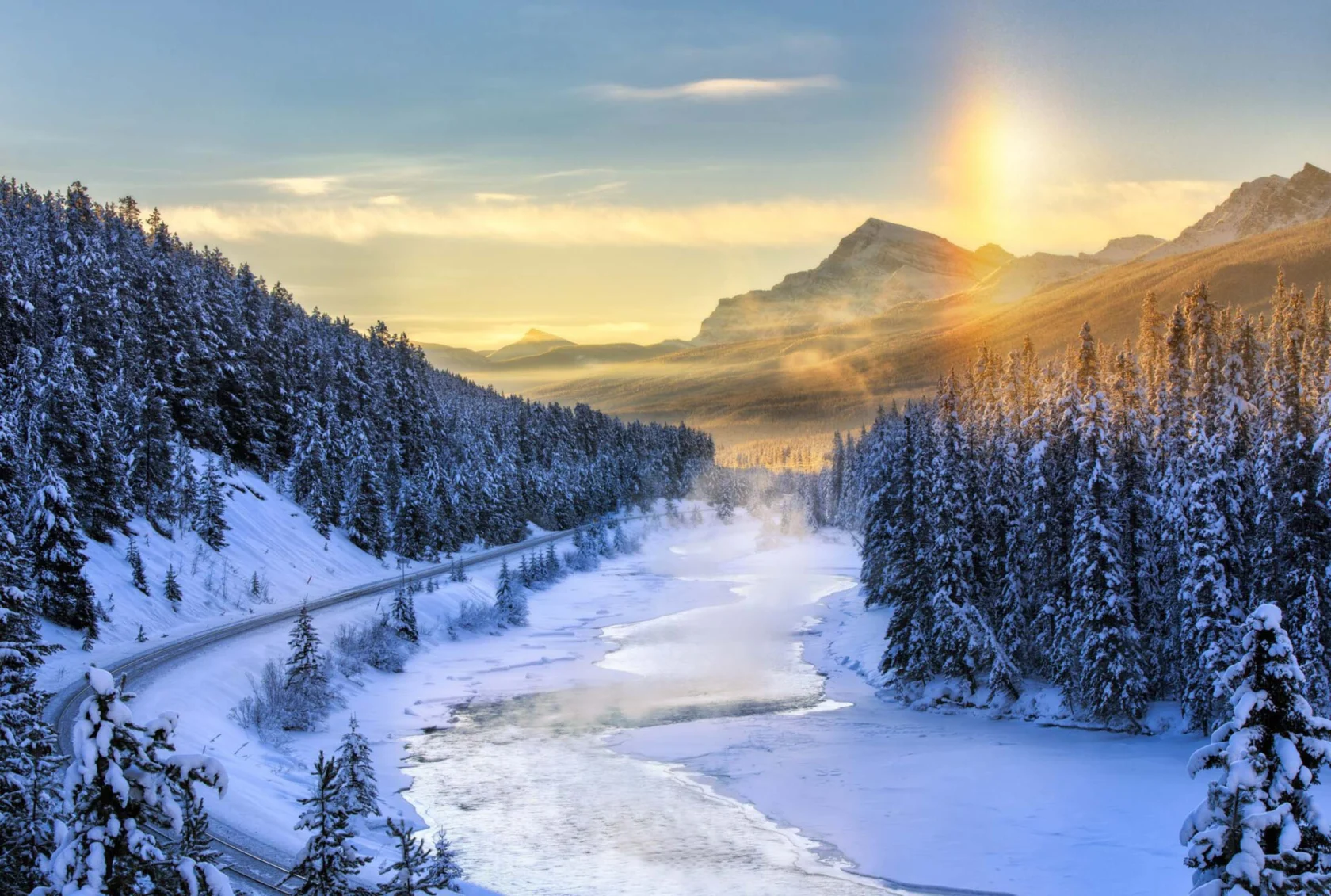 Snow-covered mountains and pine trees surround a frozen river with a winding road. The sun is low, casting a warm glow over the landscape.