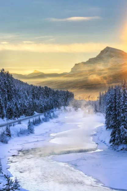 Snow-covered mountains and pine trees surround a frozen river with a winding road. The sun is low, casting a warm glow over the landscape.