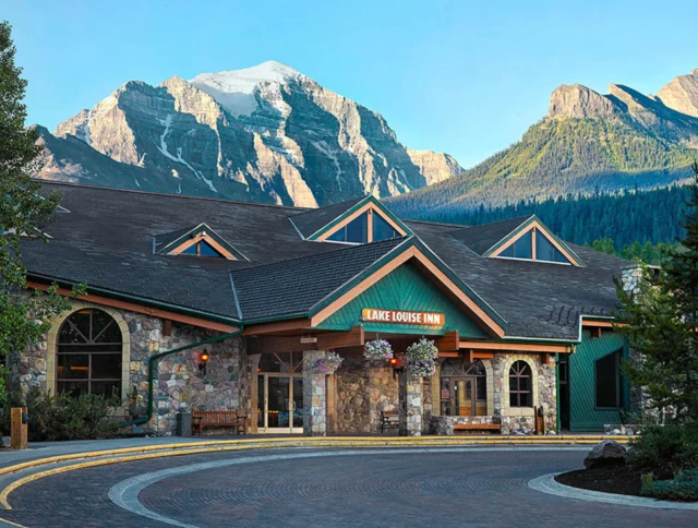 Mountain lodge with stone facade and green accents, surrounded by trees, with snow-capped peaks in the background.