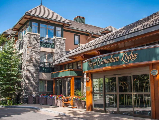 Exterior view of a lodge with stone and wood accents, featuring a sign that reads "Royal Canadian Lodge." Surrounded by trees under a clear blue sky.