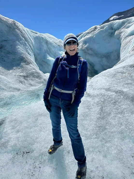 Smiling person in blue jacket and jeans standing on a glacier, wearing sunglasses, a beanie, and hiking boots. Clear blue sky in the background.