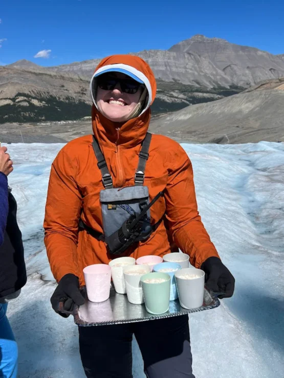 Person in orange jacket standing on a glacier holding a tray with colorful cups.