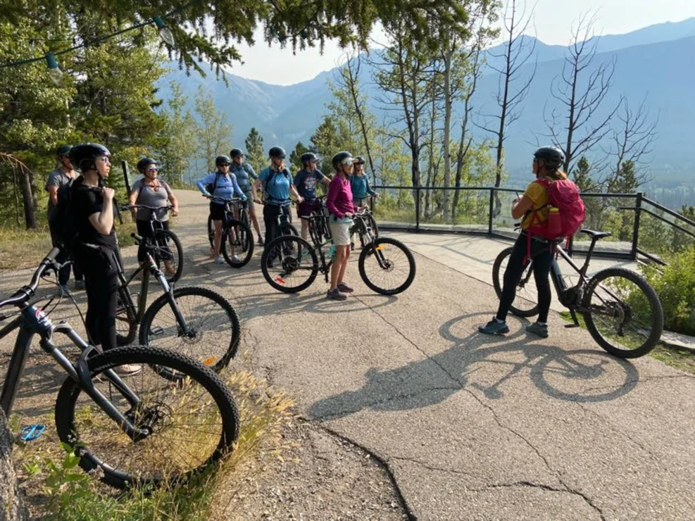 A group of people with bicycles gathers on a paved path in a forested area, with mountains visible in the background.