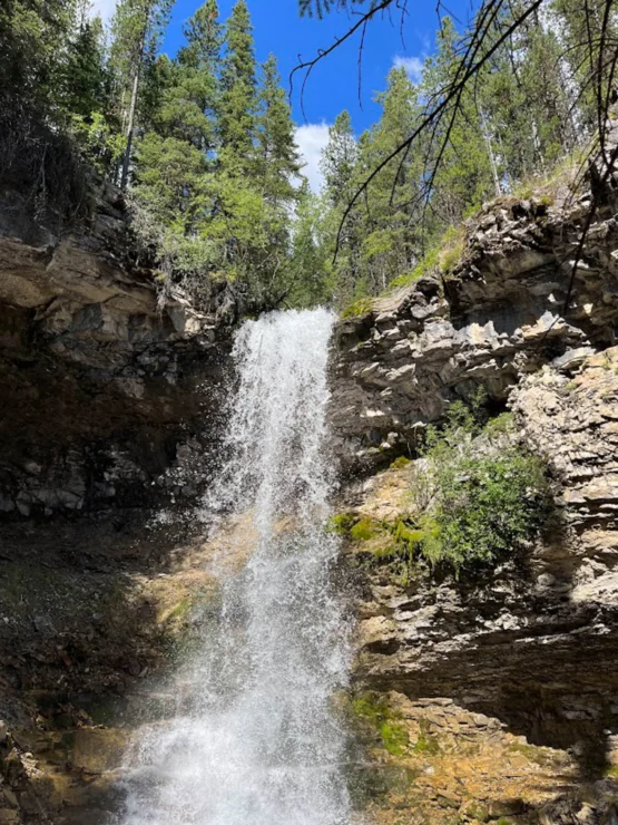 A waterfall cascades over rocky cliffs surrounded by trees, with a clear blue sky above.