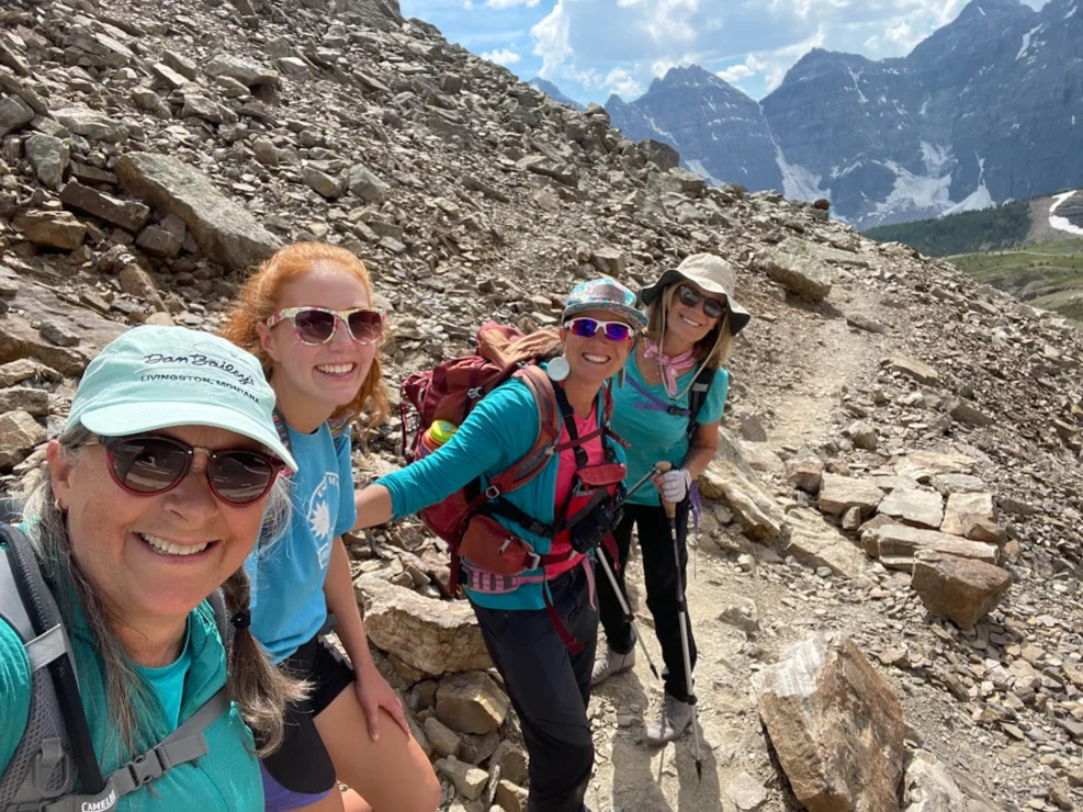 Four women hikers in turquoise shirts smile on a rocky mountain trail with rugged peaks and a partly cloudy sky in the background.