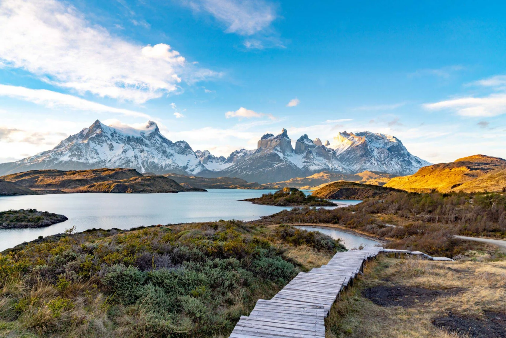 A wooden boardwalk leads to a scenic view of snowy mountains by a lake under a blue sky with scattered clouds.