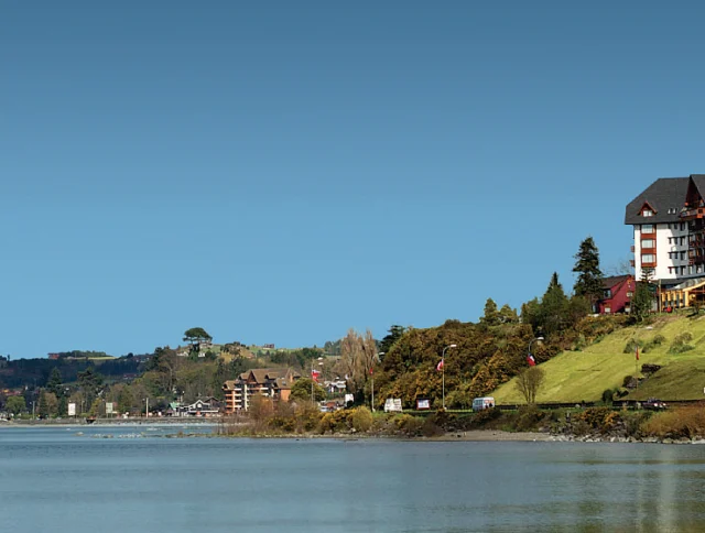 Lakeside hotel on a grassy hill with trees, buildings, and a calm body of water under a clear blue sky.