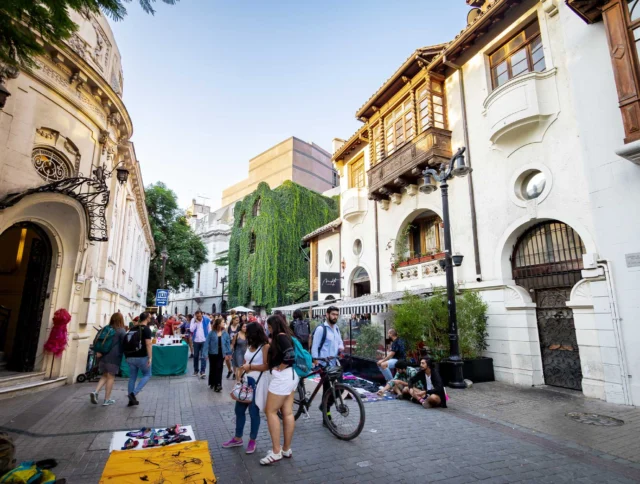 Street scene with people walking past ornate buildings and street vendors. A person sits next to a bicycle on the right, with greenery covering part of a building in the background.