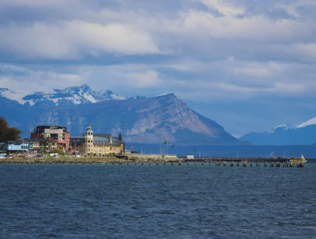 Coastal town with colorful buildings along a pier, set against a backdrop of snow-capped mountains and a cloudy sky.