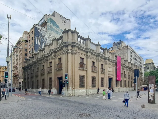 Street view of a historic building with columns and ornate trim, surrounded by pedestrians and modern city architecture. Traffic lights and signs are visible in the urban scene.