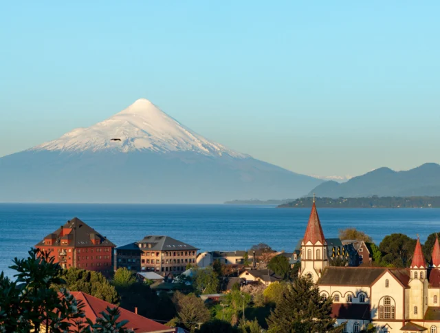 Scenic view of a town with a church and buildings near a lake, with a snow-capped mountain in the background under a clear blue sky.