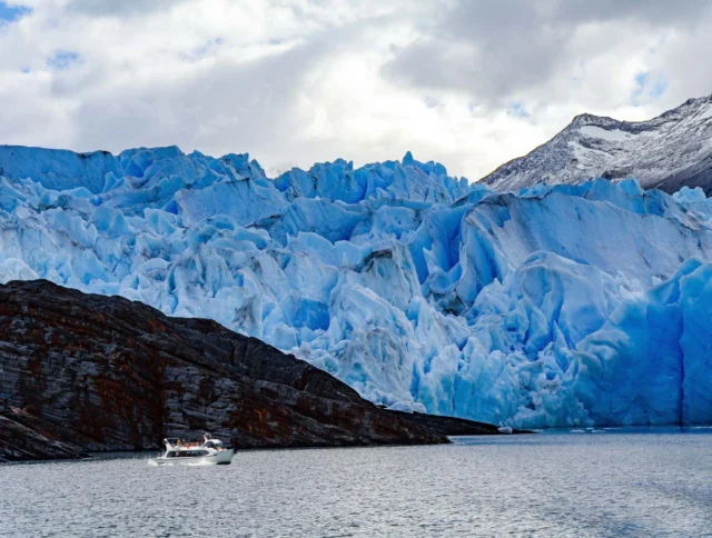 A small boat navigates near a massive glacier with jagged ice formations under a cloudy sky, with a rocky shoreline visible in the foreground.