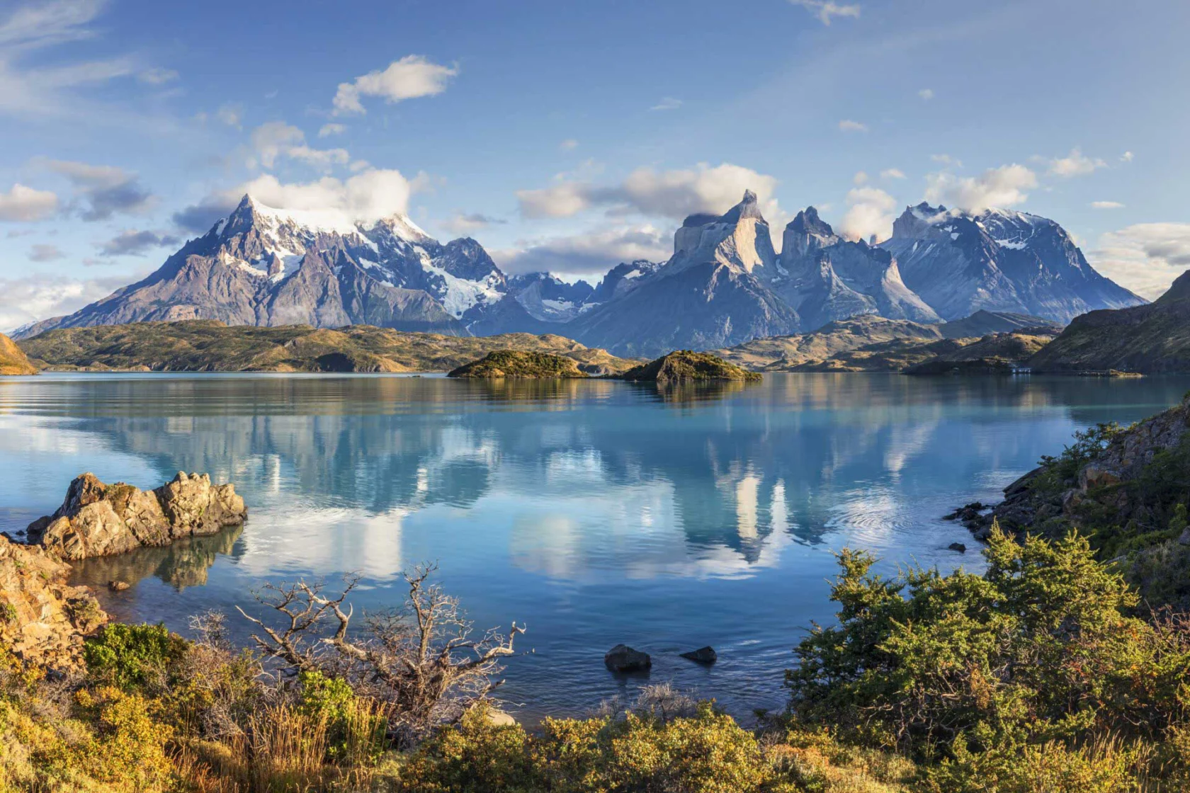 A serene lake with crystal-clear water reflecting snow-capped mountains and a clear blue sky, surrounded by lush greenery and rocky terrain.