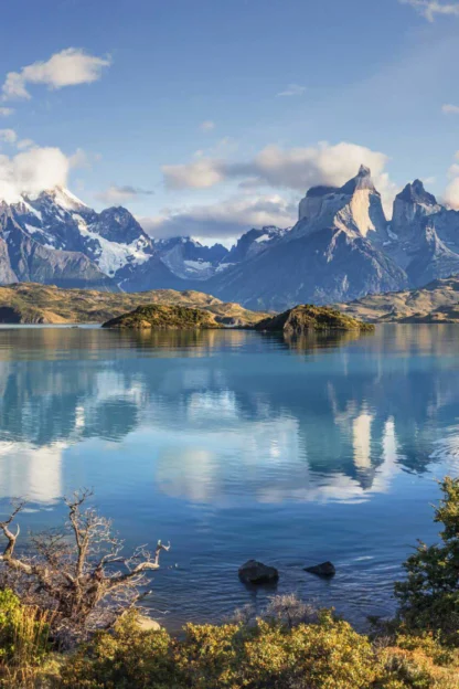 A serene lake with crystal-clear water reflecting snow-capped mountains and a clear blue sky, surrounded by lush greenery and rocky terrain.