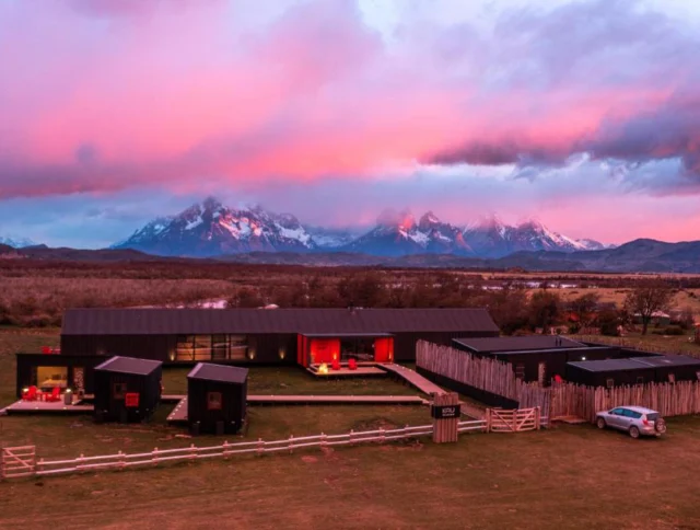 Modern black house with large windows, set against a dramatic pink and purple sky, with mountains in the background. A car is parked in the foreground on a grassy field.