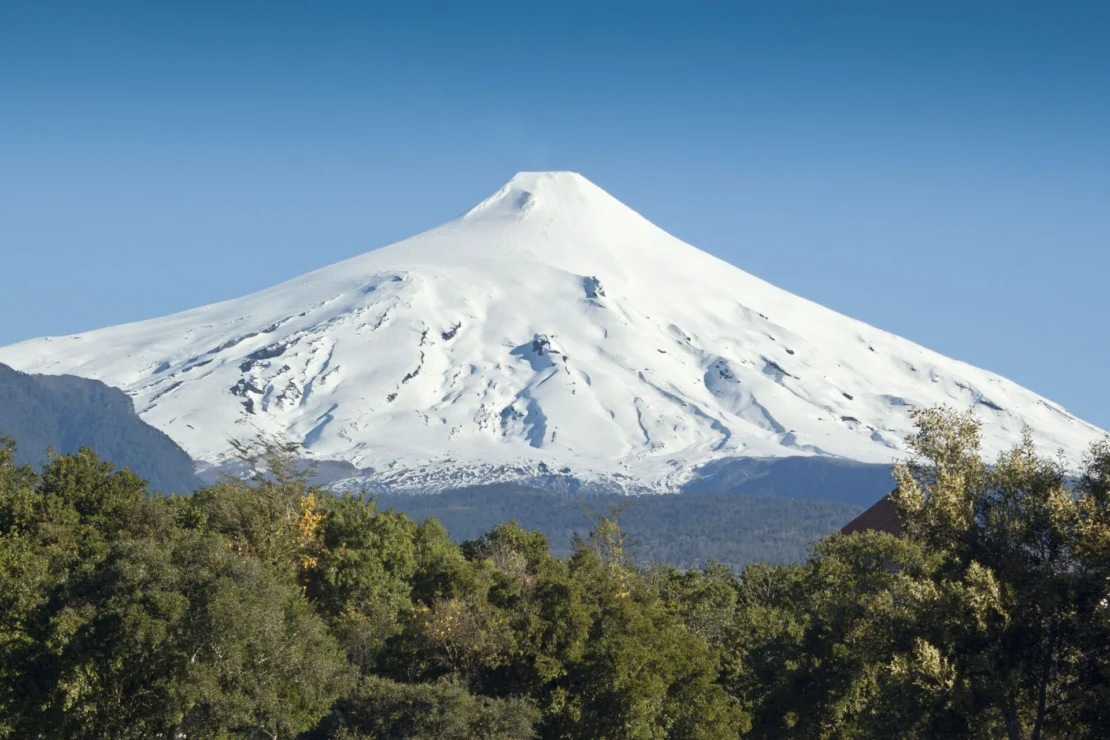 Snow-covered volcano under a clear blue sky, surrounded by green trees.